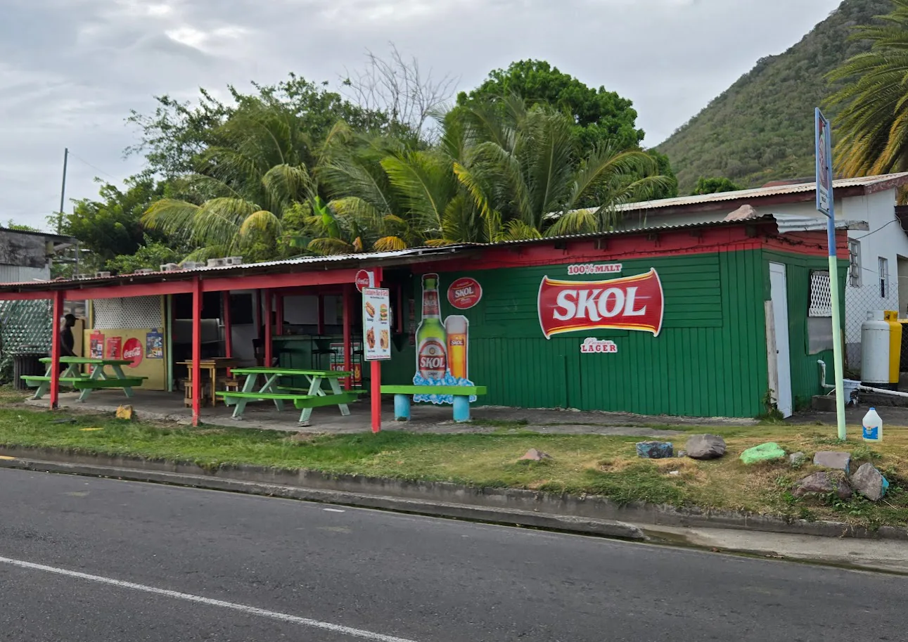 A green roadside bar with red posts and picnic tables, backed by palms and a green hillside.