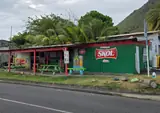 A green roadside bar with red posts and picnic tables, backed by palms and a green hillside.