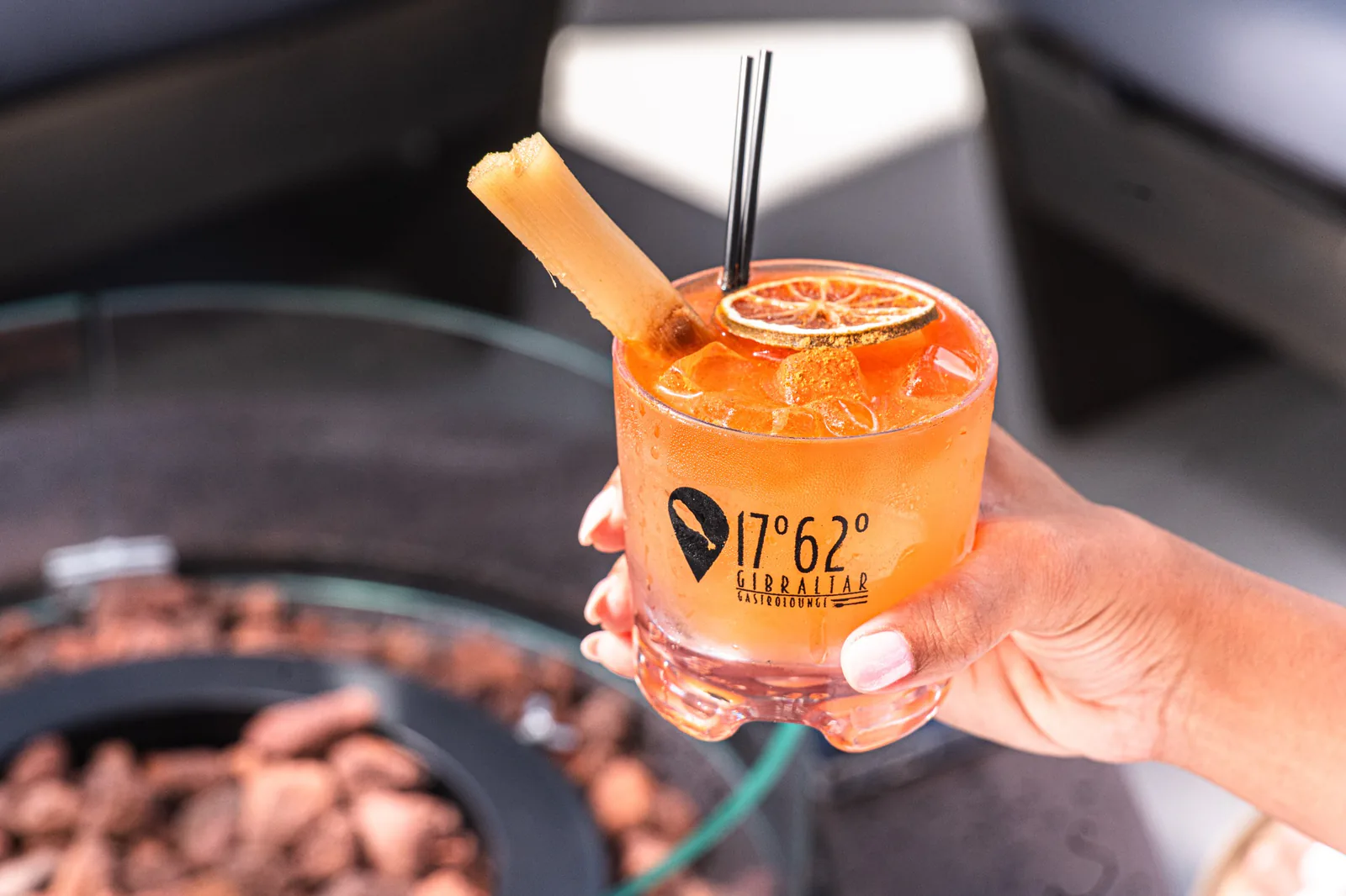 An orange drink with a citrus garnish in a Gibraltar Gastrolounge glass, held beside the lounge seating.